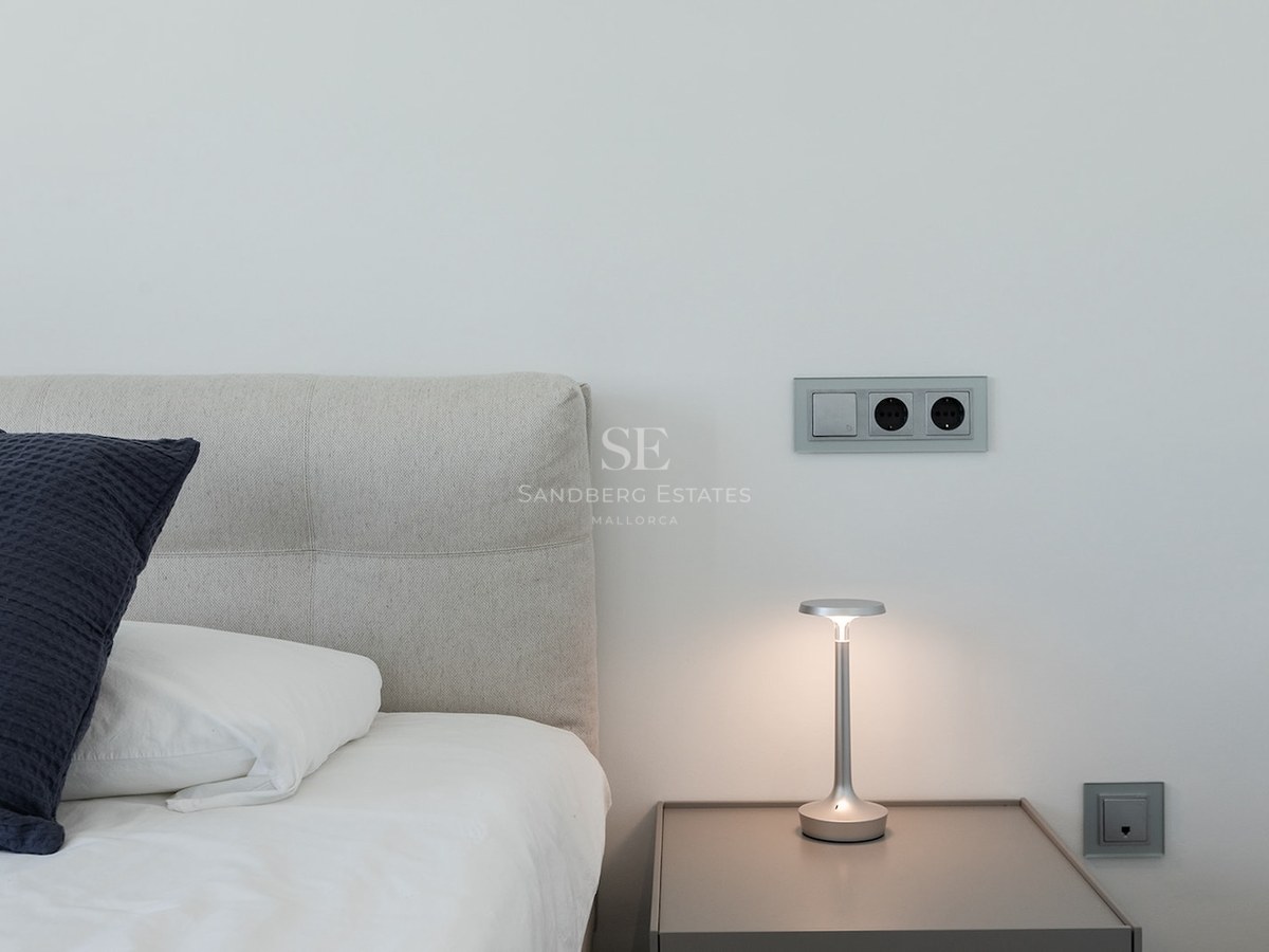 Close-up of a bed with grey headboard, nightstand with a designer lamp, and modern electrical sockets on a white wall.