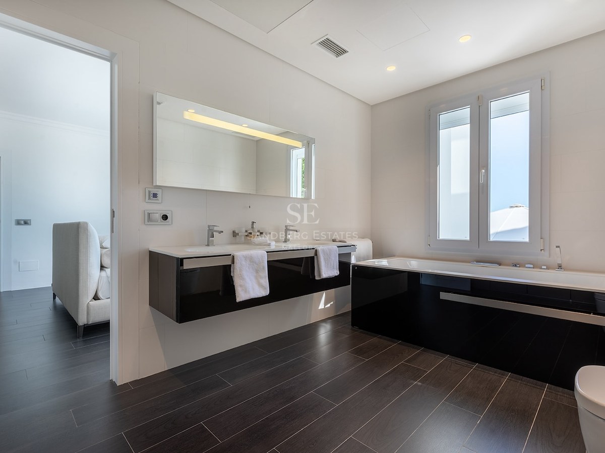Modern bathroom with double vanity, black bathtub, and dark flooring next to a bright bedroom.