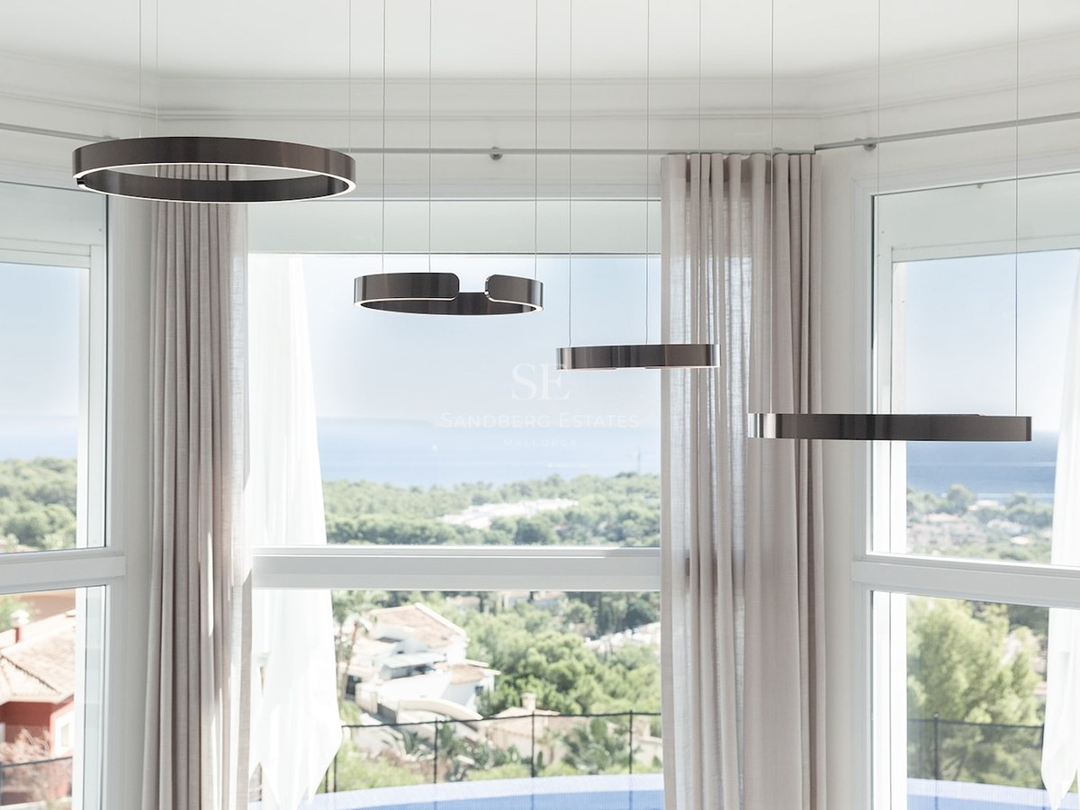 Four bronze ring-shaped pendant lights hanging in front of a large bay window overlooking a coastal landscape and the sea.
