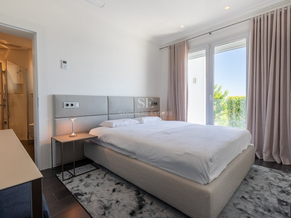 Bright master bedroom with a grey upholstered bed, white linens, ensuite bathroom, and large windows looking onto greenery.