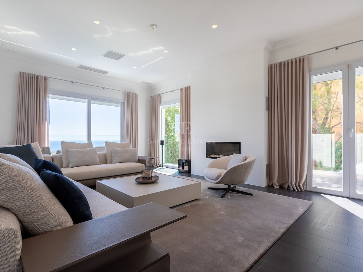 Spacious living room with cream sofas, built-in fireplace, dark flooring, and large windows overlooking the sea.