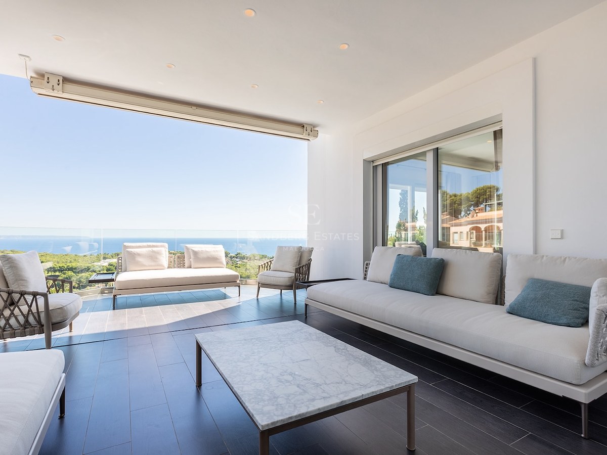 Modern covered terrace with white sofas, a marble coffee table, and a glass railing overlooking the Mediterranean sea.