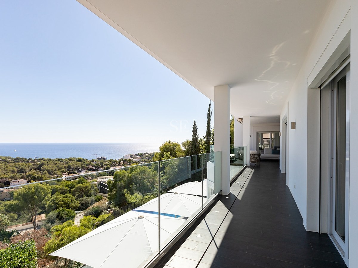 Spacious white balcony with glass railing overlooking a Mediterranean forest and the blue ocean.