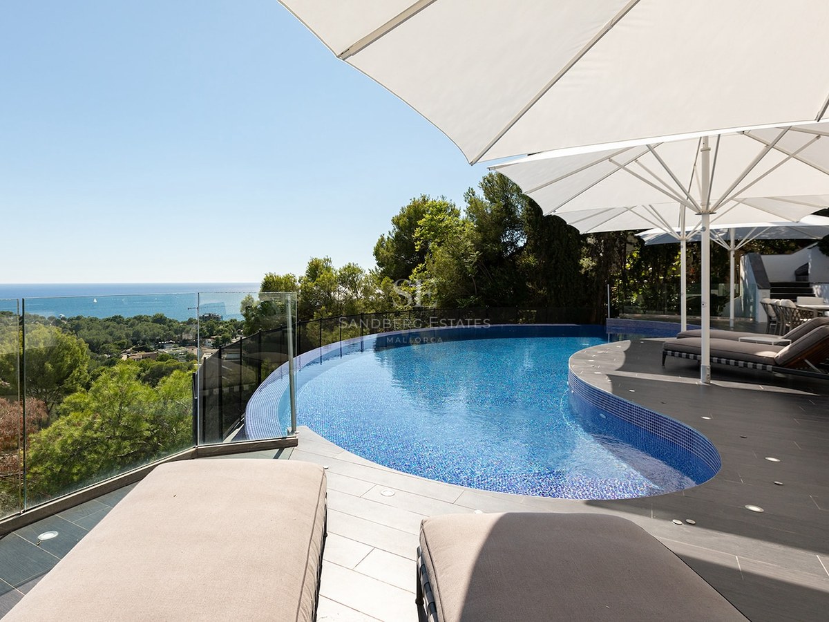 A curved blue swimming pool on a modern stone terrace with white umbrellas and sunbeds overlooking the sea.