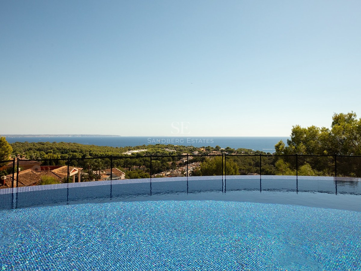 View from a blue mosaic infinity pool looking out over green trees towards the Mediterranean sea under a clear sky.