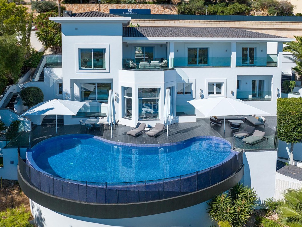 Elevated view of a modern white villa featuring a large blue semicircular infinity pool and furnished terraces.