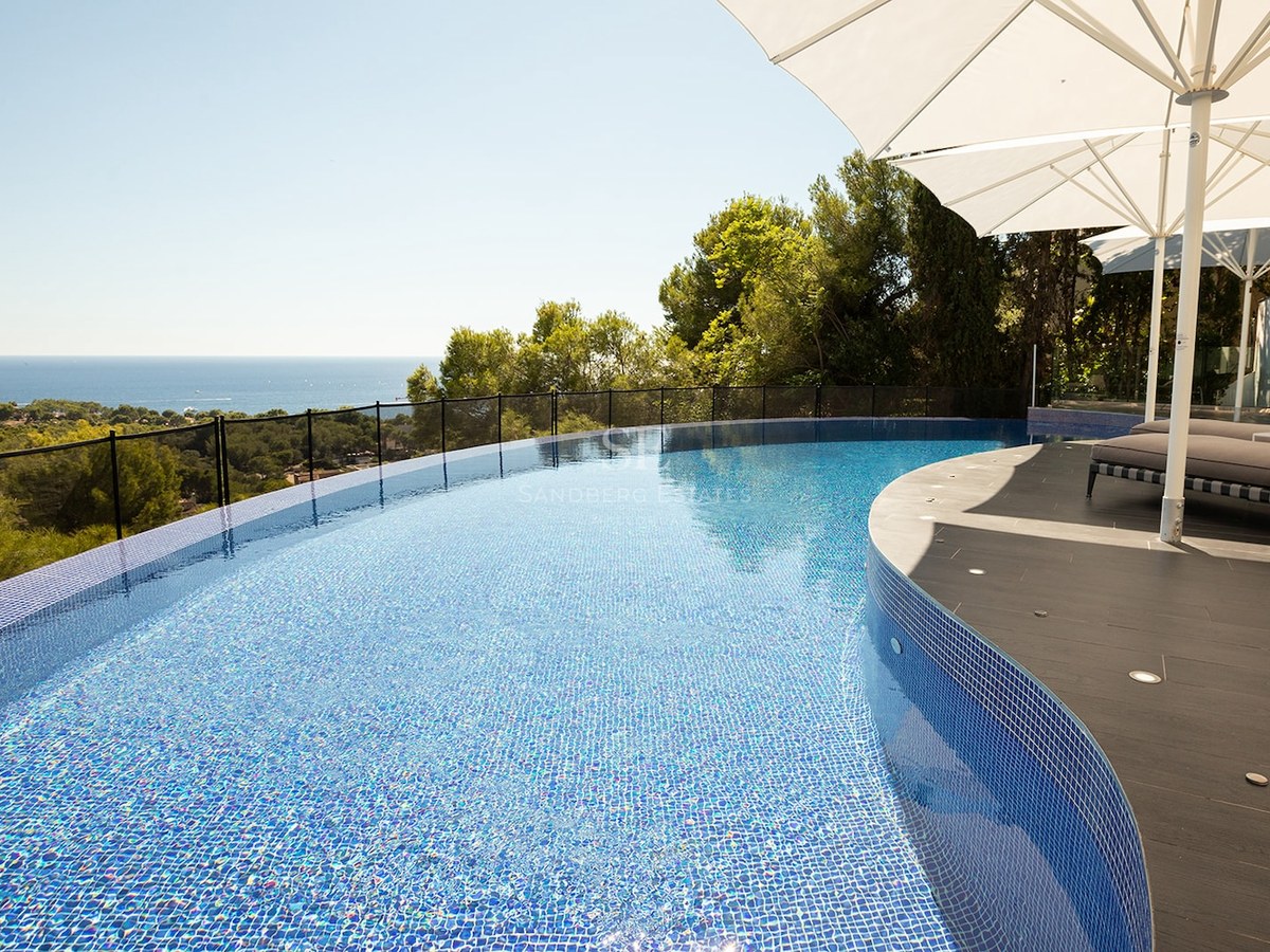 Curved infinity pool with blue mosaic tiles overlooking a lush green landscape and the blue ocean under a clear sky.