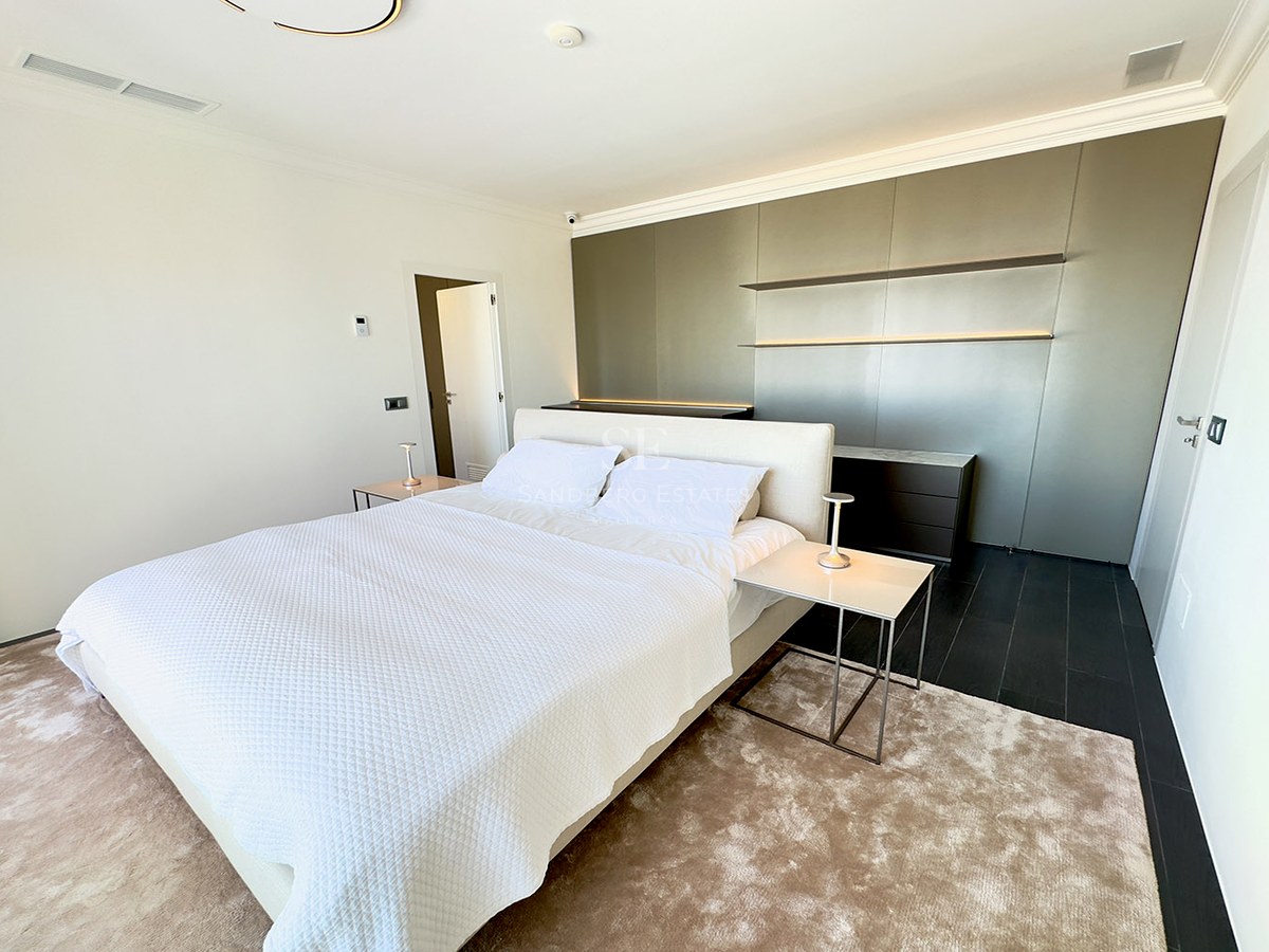 Modern bedroom featuring a large white bed, minimalist nightstands, a taupe feature wall, and a plush beige rug.
