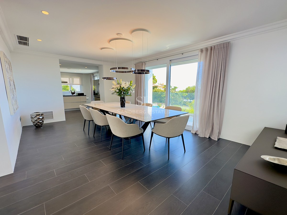 Minimalist dining room featuring a marble table, six beige chairs, dark flooring, and large windows viewing a garden.
