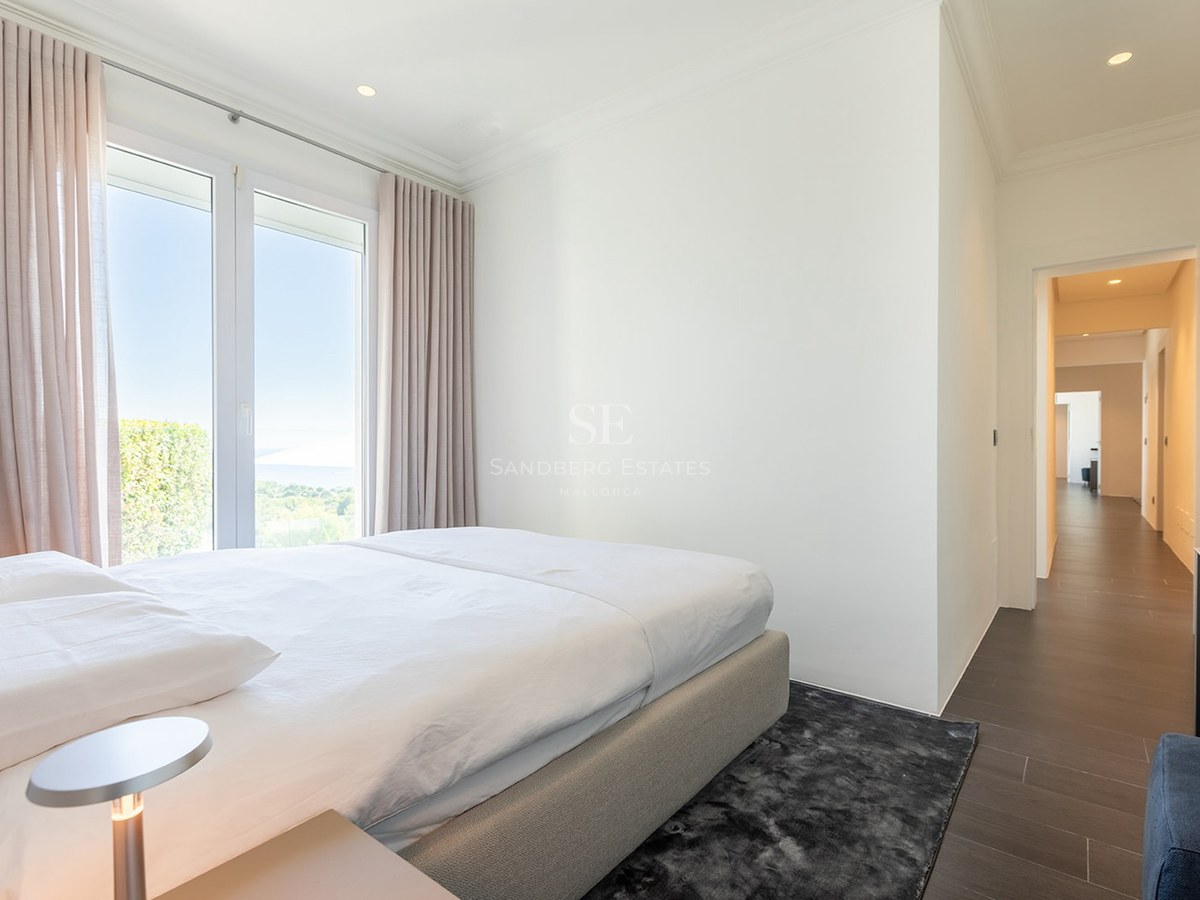 Bright bedroom featuring a white bed, dark rug, and large window overlooking the sea and lush greenery.