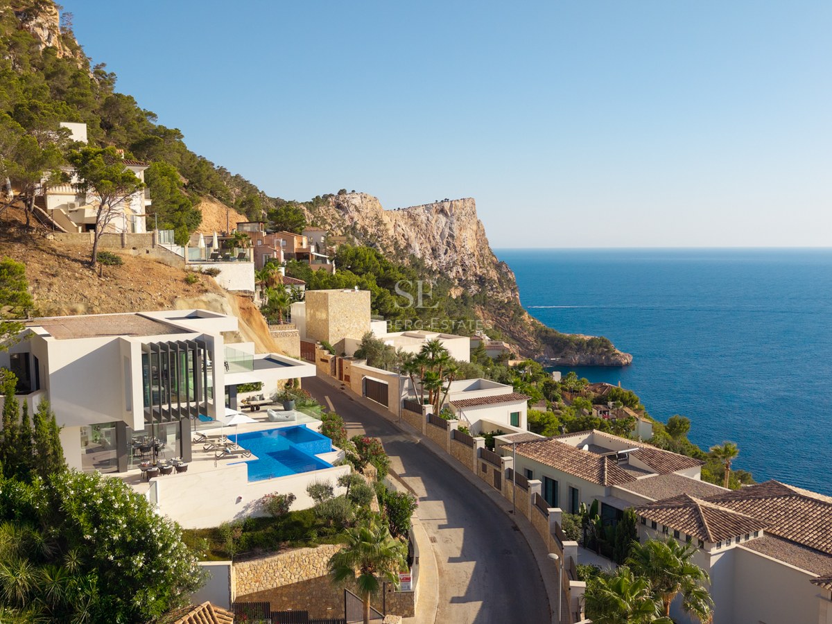 Aerial view of a white modern villa with an infinity pool on a cliffside overlooking the blue Mediterranean sea.