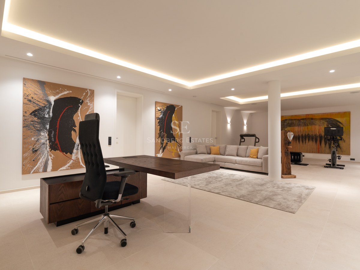 Spacious modern home office with a dark wood desk, grey sofa, abstract art, and gym equipment in the background.