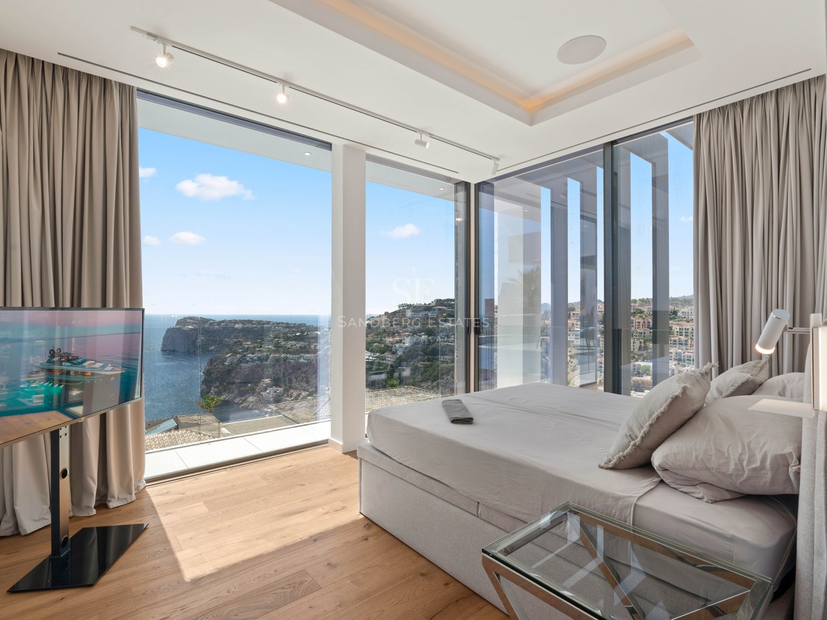 Modern bedroom with floor-to-ceiling windows showing a panoramic view of the sea and coastline.