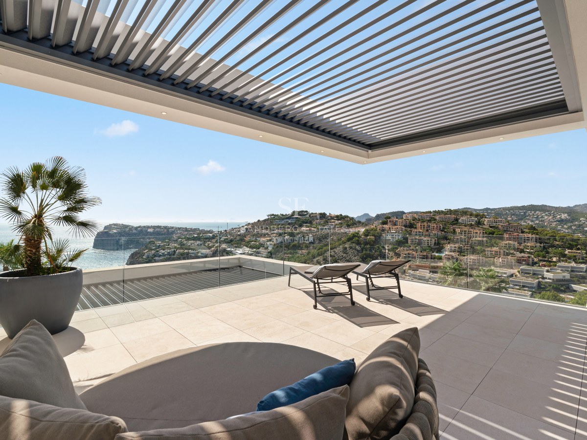 Modern terrace with stone tiling, sun loungers, a louvered pergola, and stunning views of the sea and coastal hills.