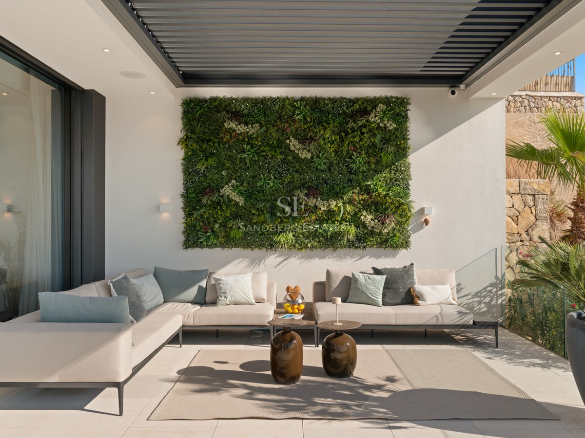 A contemporary outdoor terrace featuring a large beige corner sofa, a lush green vertical garden wall, and a louvered pergola.