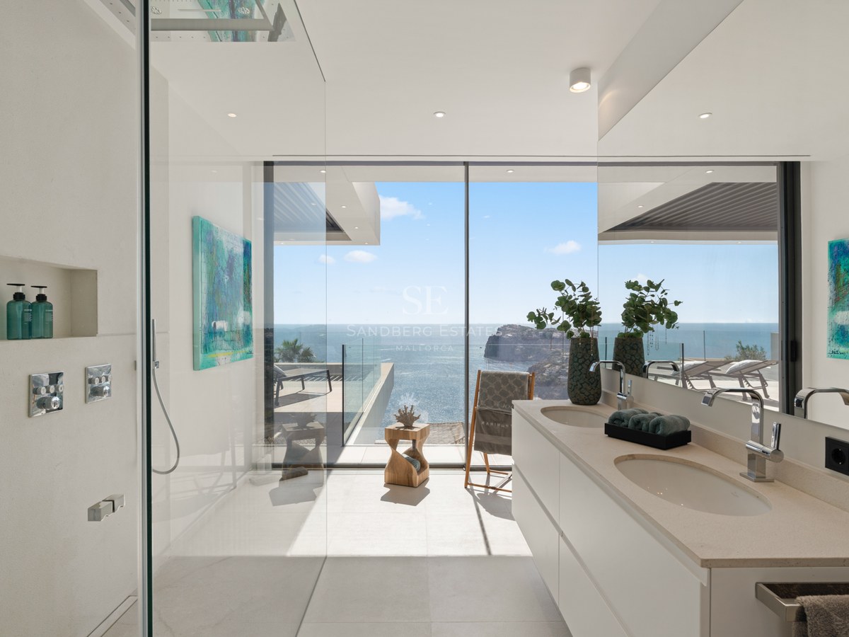 Modern white bathroom with glass walk-in shower and double vanity overlooking the ocean through floor-to-ceiling windows.