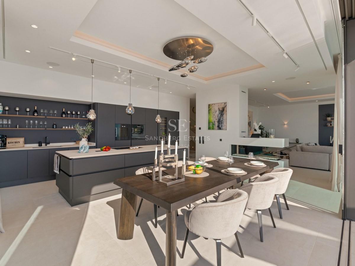 Modern dining area with dark wood table, beige chairs, and sleek gray kitchen featuring floor-to-ceiling windows.