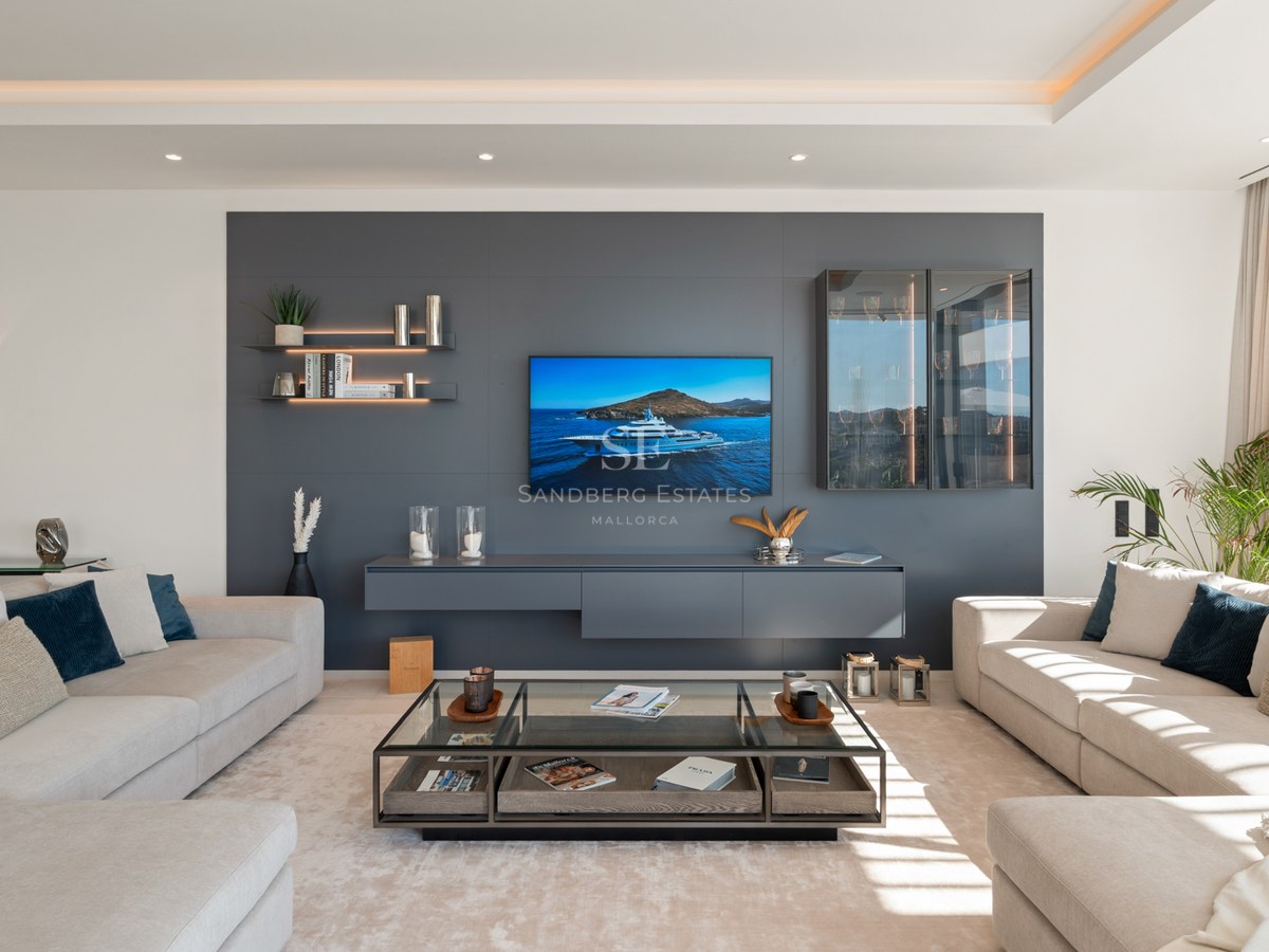 A bright, modern living room with cream sofas, a large grey media wall with TV, and a glass coffee table.