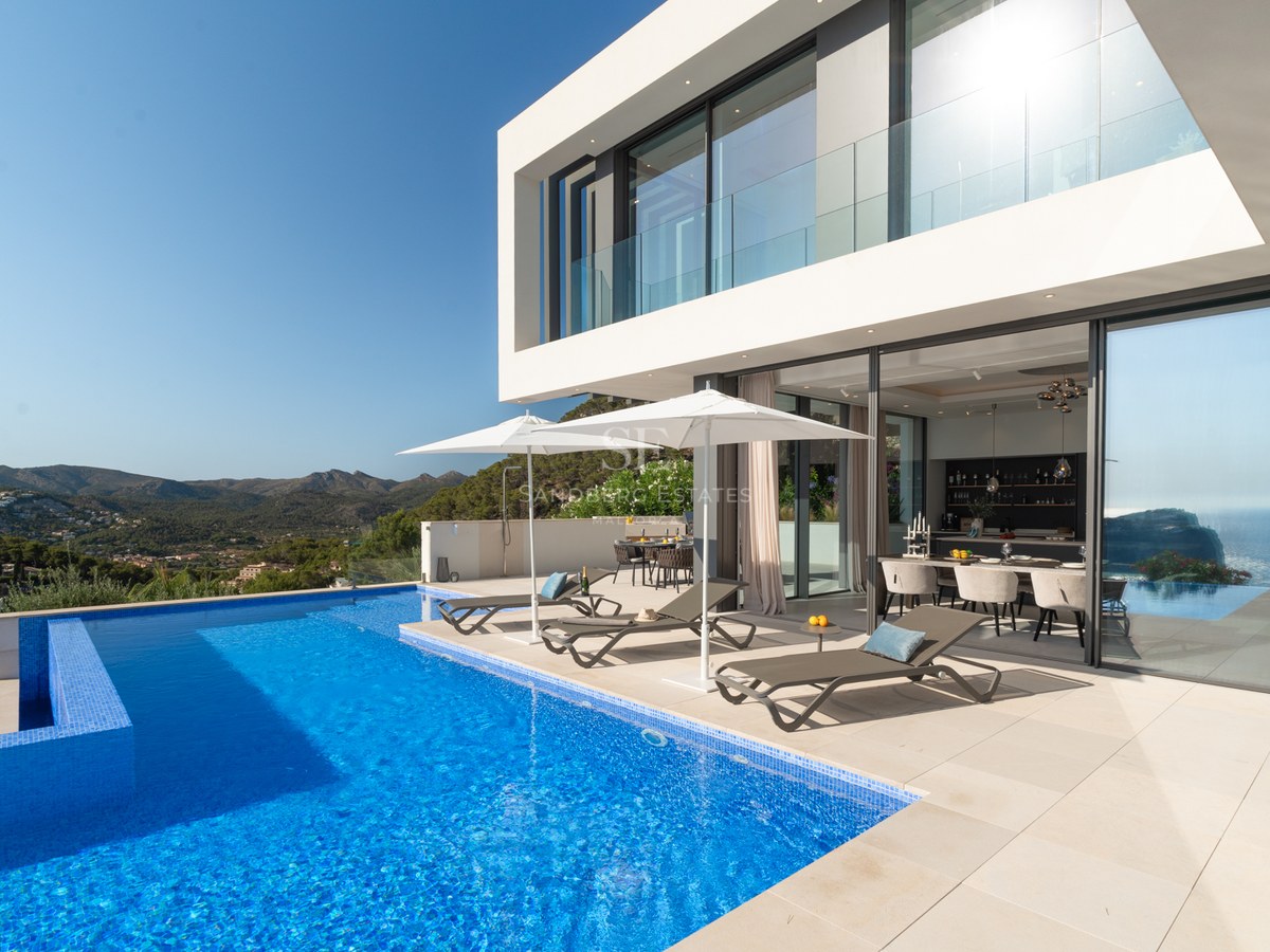 Luxury infinity pool and white modern villa with mountain views and designer loungers under white umbrellas.