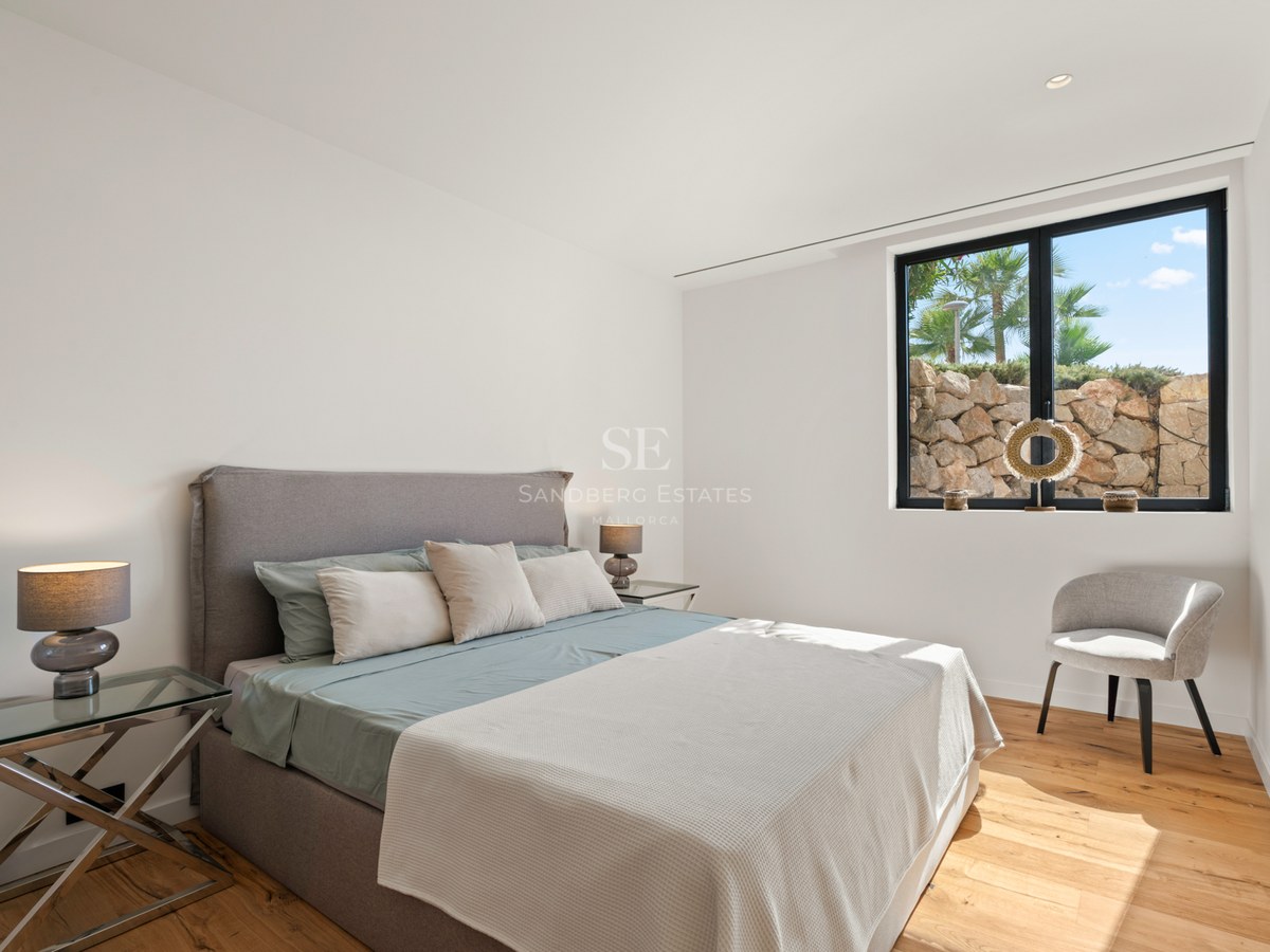 Bright modern bedroom with a gray upholstered bed, glass nightstands, wooden floors, and a large window.