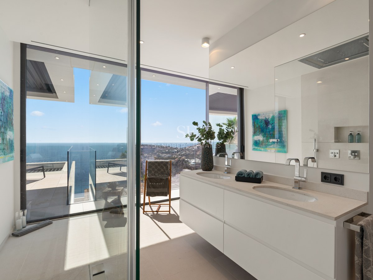 Modern bathroom featuring a double white vanity and floor-to-ceiling windows overlooking the Mediterranean sea and coastline.
