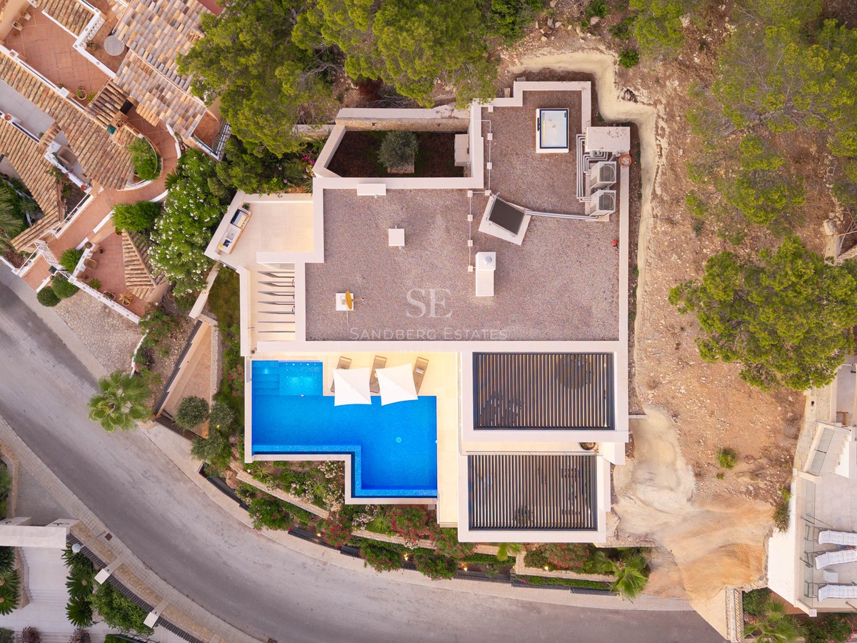 Flygvy uppifrån av en modern vit villa med en klarblå L-formad pool och solstolar.