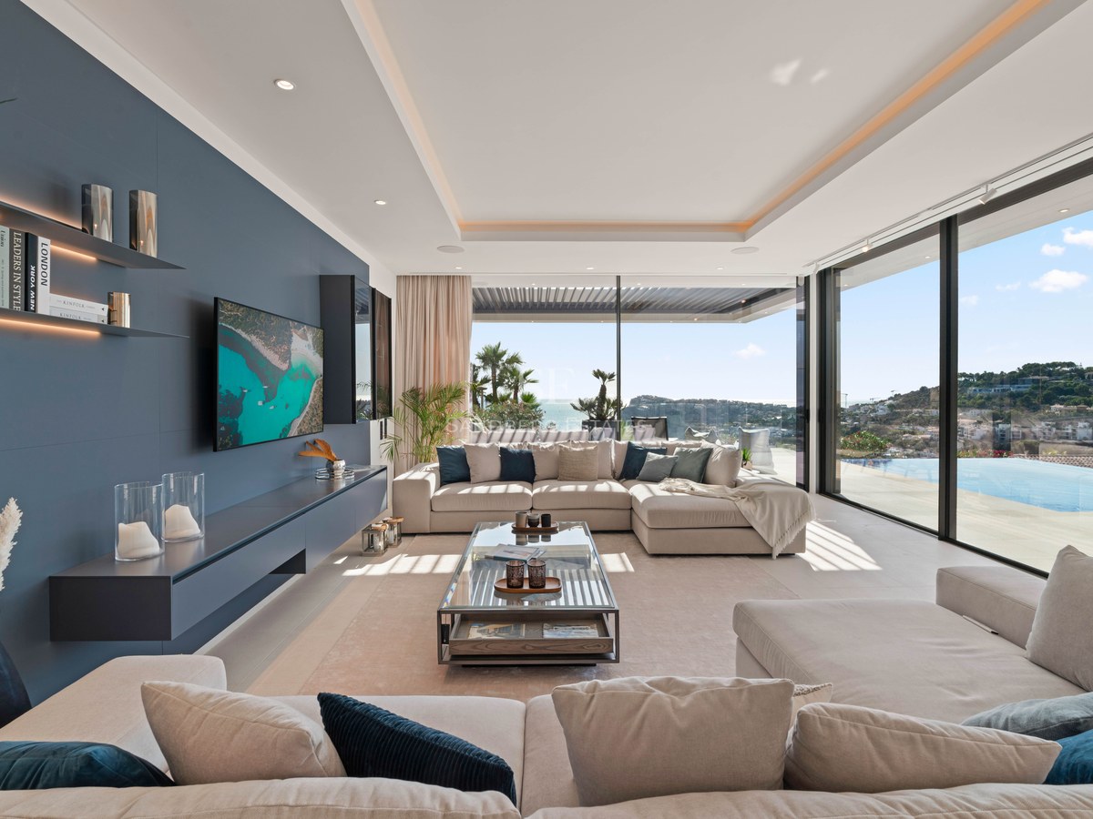Spacious modern living room with beige sofas, a blue accent wall, and large windows overlooking the sea and an infinity pool.