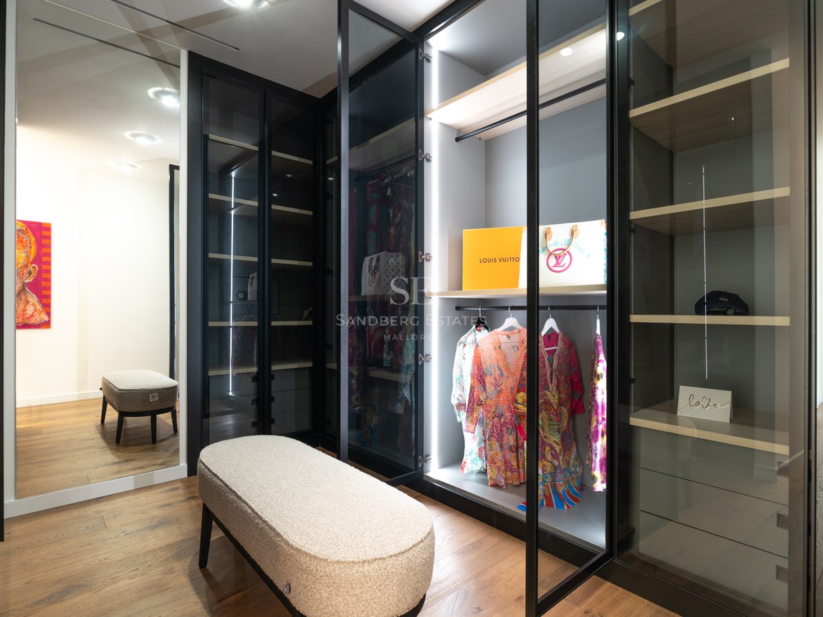 Modern walk-in closet featuring glass-door wardrobes with LED lighting, wood floors, and a textured boucle bench.