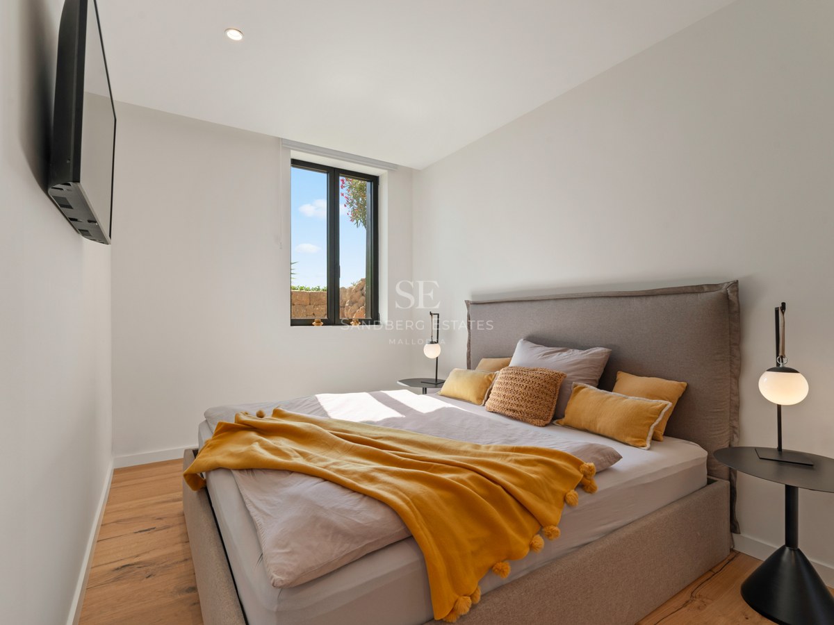 Modern bedroom with a grey bed, mustard yellow throw, wooden floors, and a window showing a bright sky.