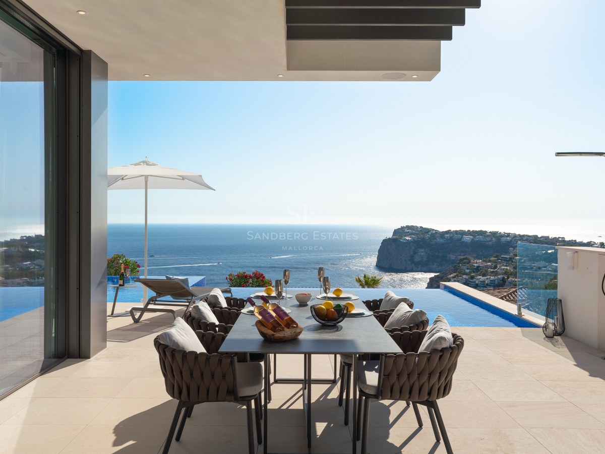 Modern outdoor terrace with a dining table overlooking an infinity pool and a vast blue ocean with coastal cliffs.