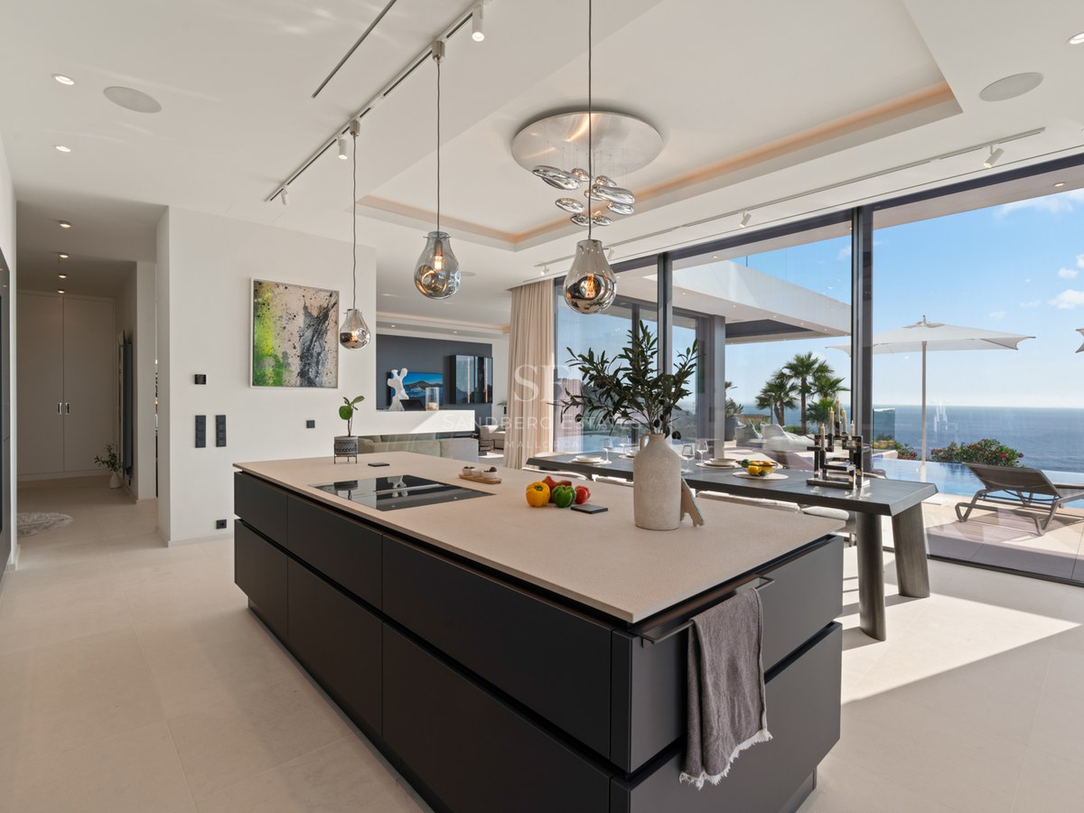 Modern kitchen with a large dark island, designer glass pendants, and floor-to-ceiling windows overlooking the ocean.