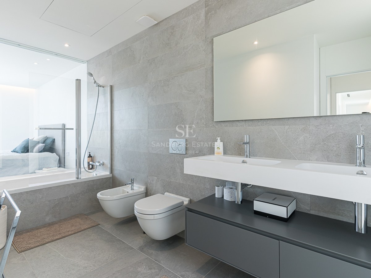 Modern bathroom featuring grey stone tiles, double vanity, wall-hung toilet, and a glass wall looking into a bedroom.