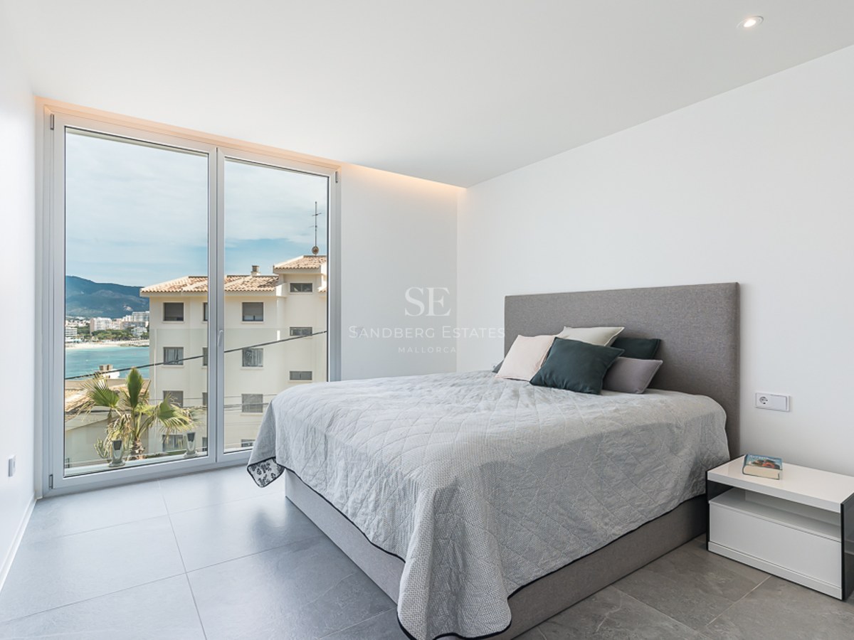 Bright modern bedroom with a large window overlooking a coastal town and sea, featuring a grey bed and tiled floor.