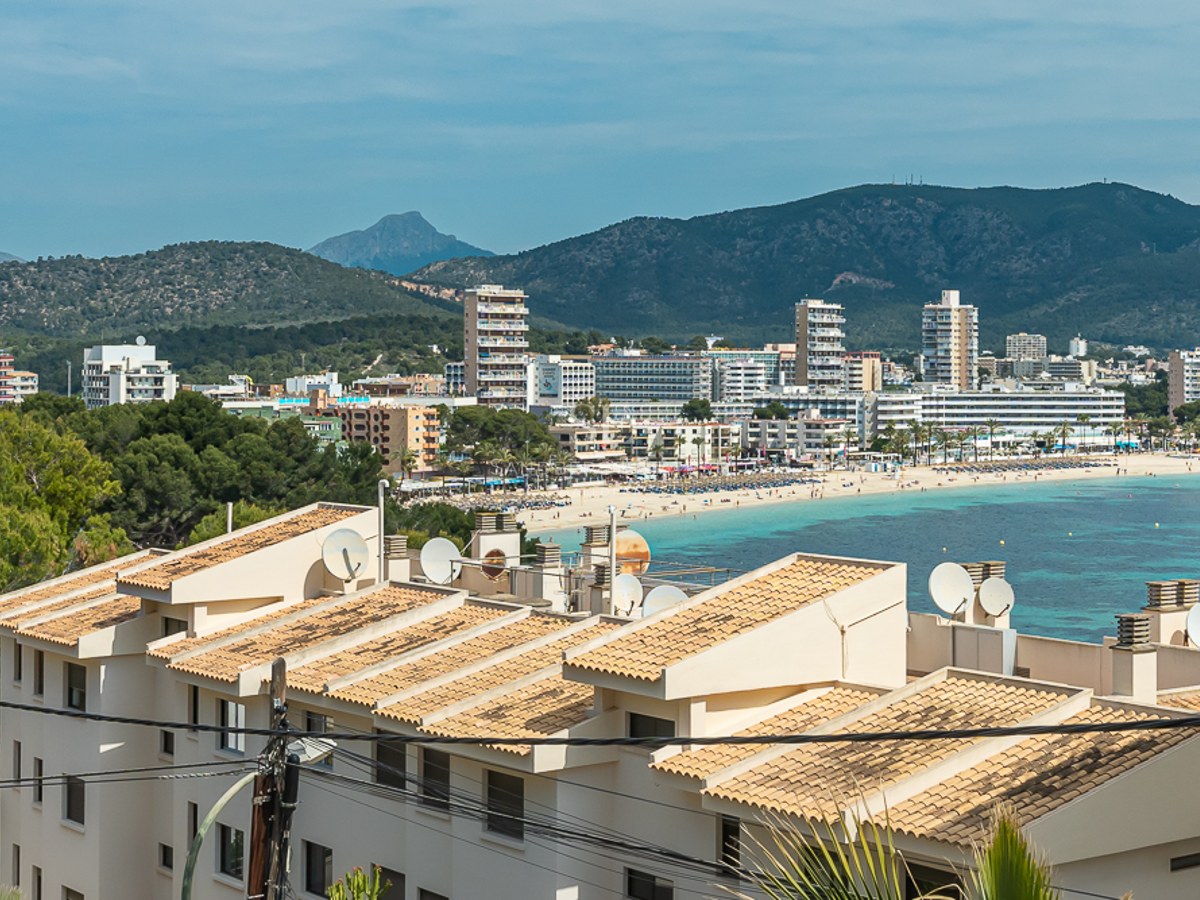 View overlooking a coastal bay with turquoise water, a sandy beach, white hotels, and green mountains in the background.