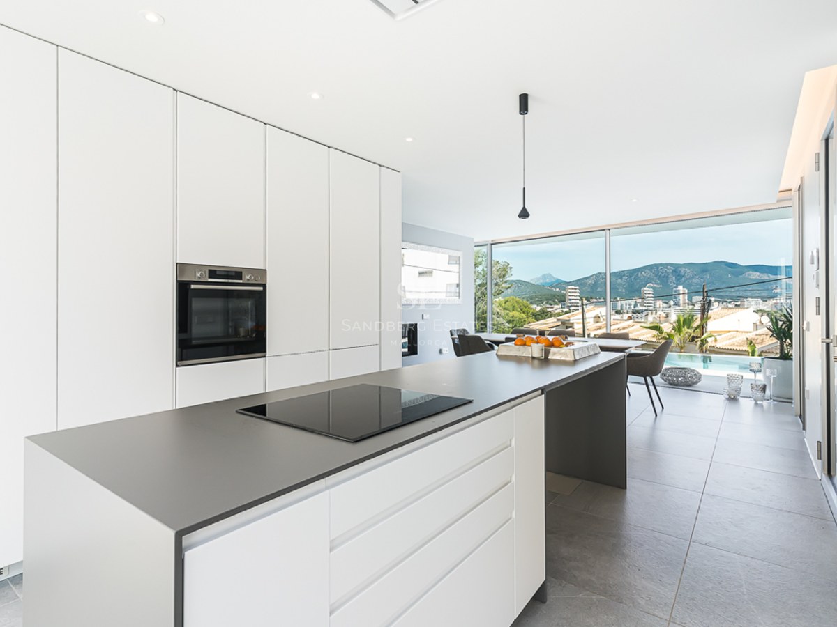 Bright white kitchen with a grey island, induction hob, and large windows overlooking mountains and a pool.