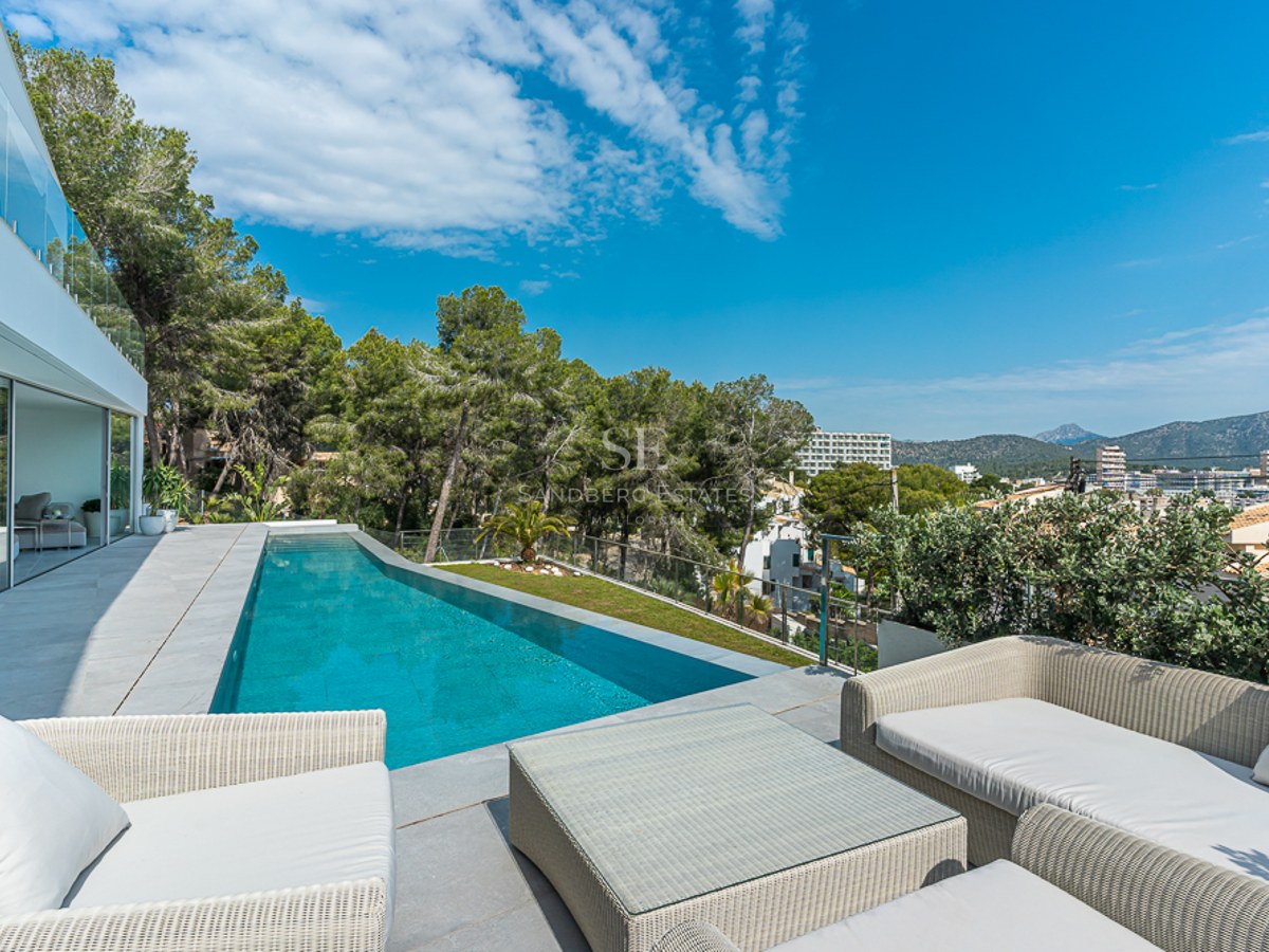 Modern terrace with a long blue swimming pool, white wicker lounge furniture, glass railings, and forest views.