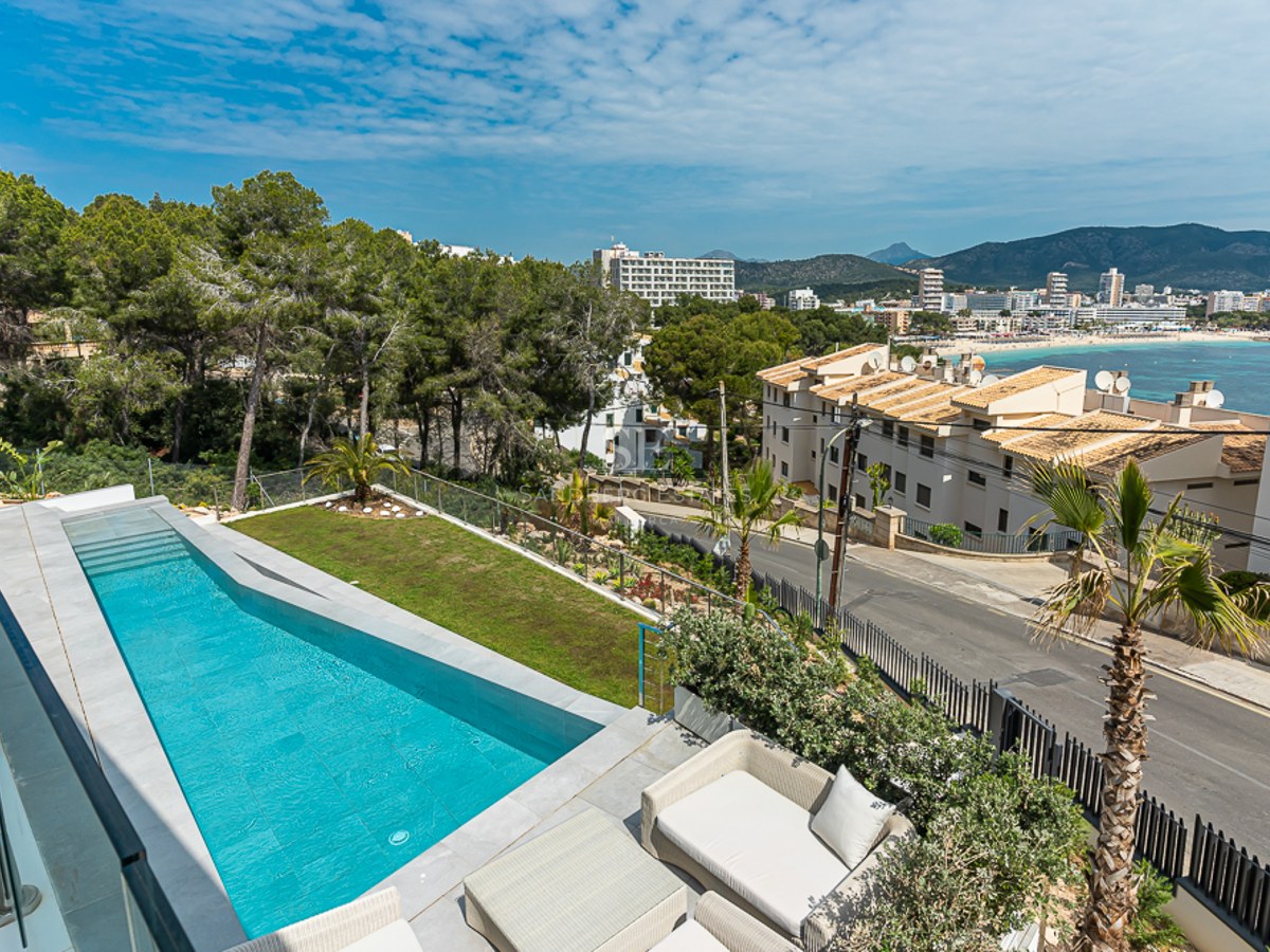 A long turquoise swimming pool on a terrace with glass railings, overlooking a coastal town and distant mountains.