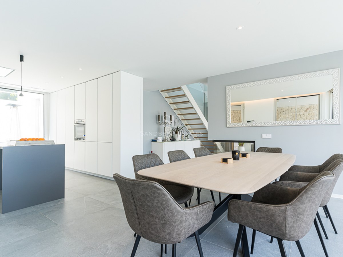 Bright dining area featuring a light wood table, grey chairs, and a minimalist white kitchen in the background.