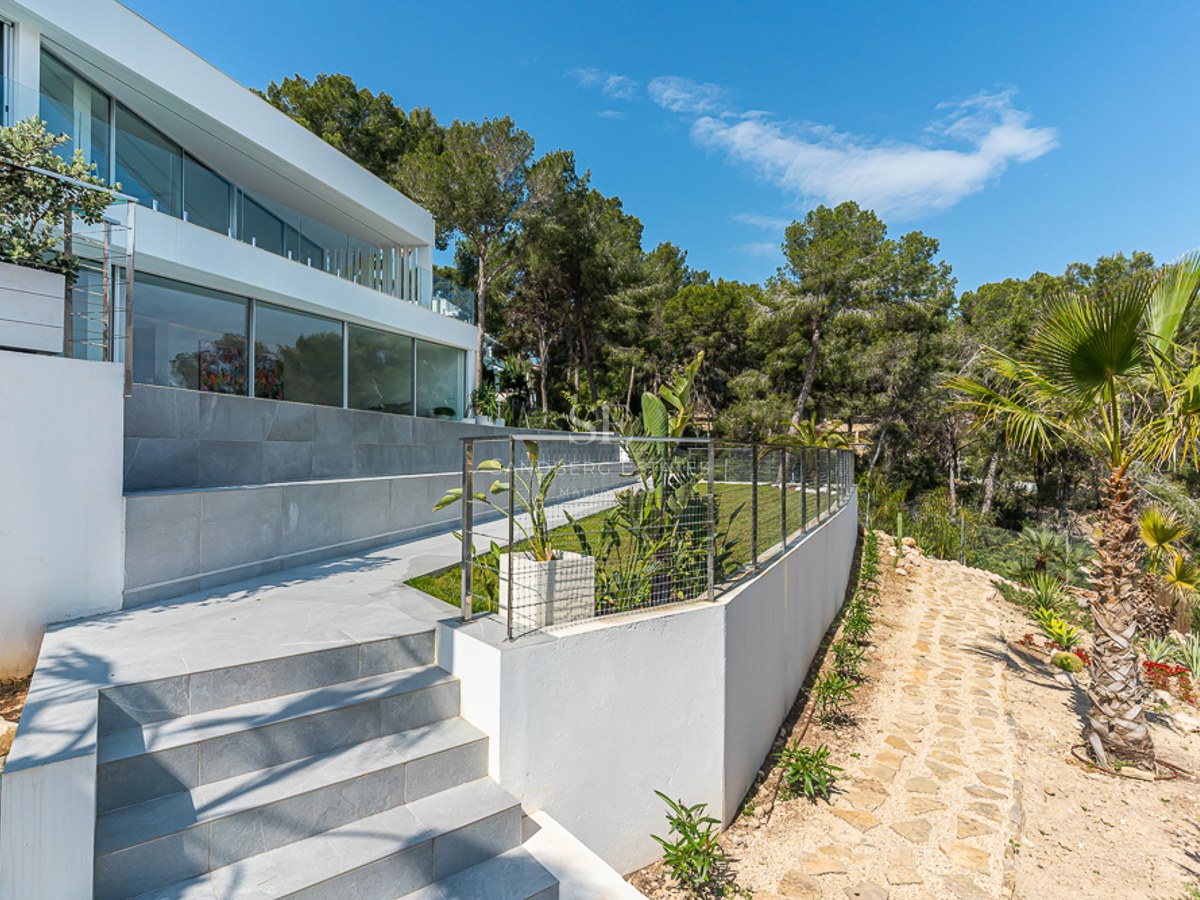 Modern white villa with a tiered garden featuring stone stairs, a lawn, and palm trees under a clear blue sky.