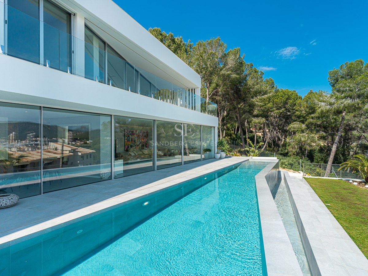 A sleek modern white villa with a long turquoise lap pool, floor-to-ceiling glass windows, and lush green trees.