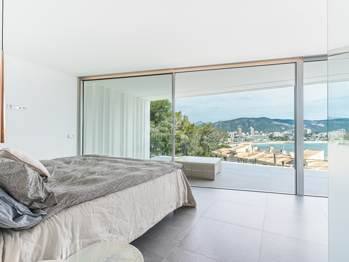 Bright modern bedroom with a large bed and floor-to-ceiling glass doors opening to a terrace with coastal views.