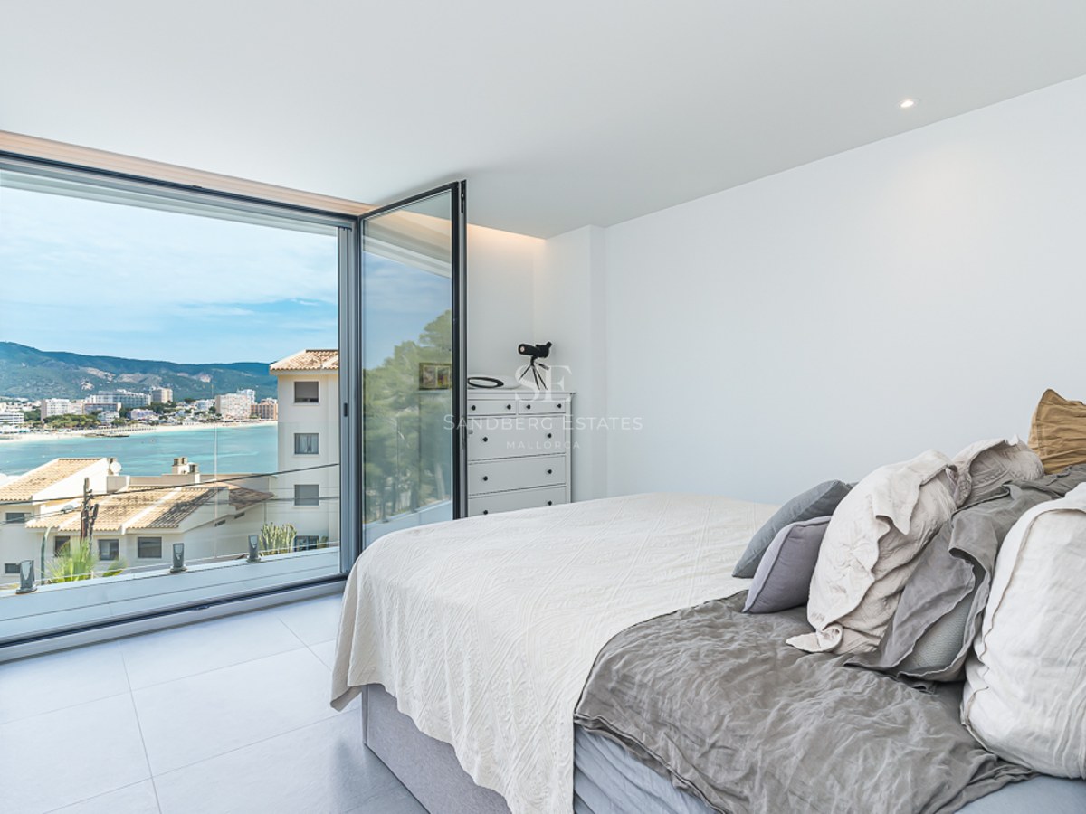 Bright bedroom with a bed and large glass doors opening to a coastal bay view and mountains.