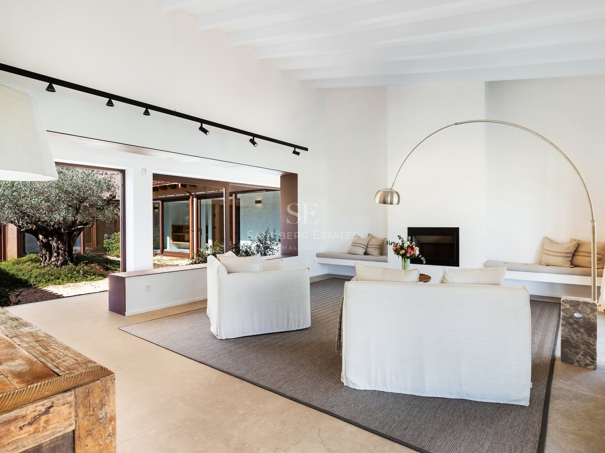 Bright living room with white sofas, an arched floor lamp, a fireplace, and views of an olive tree in a courtyard.