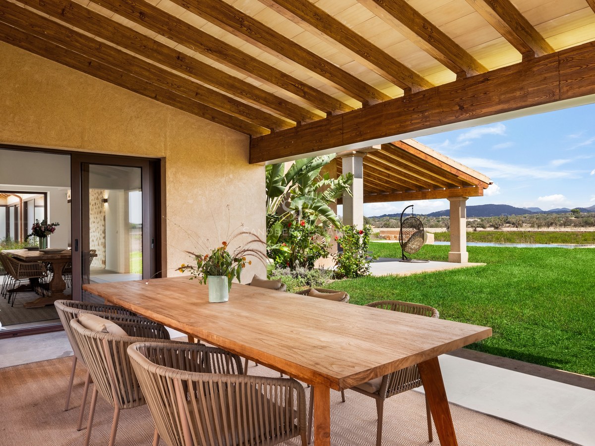 Covered terrace with a large wooden dining table and rope chairs overlooking a bright green lawn and distant hills.