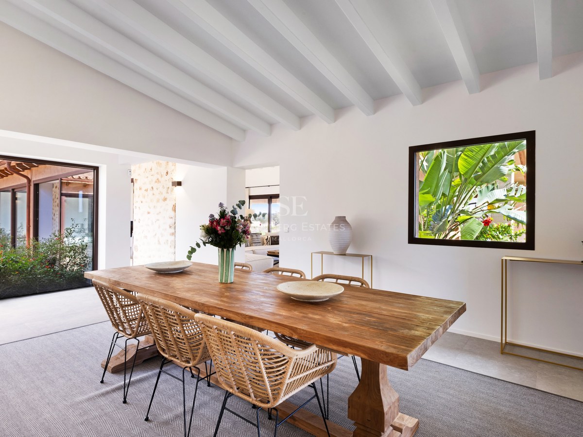 Bright dining room with a large wooden table, wicker chairs, white beamed ceiling, and lush garden views.