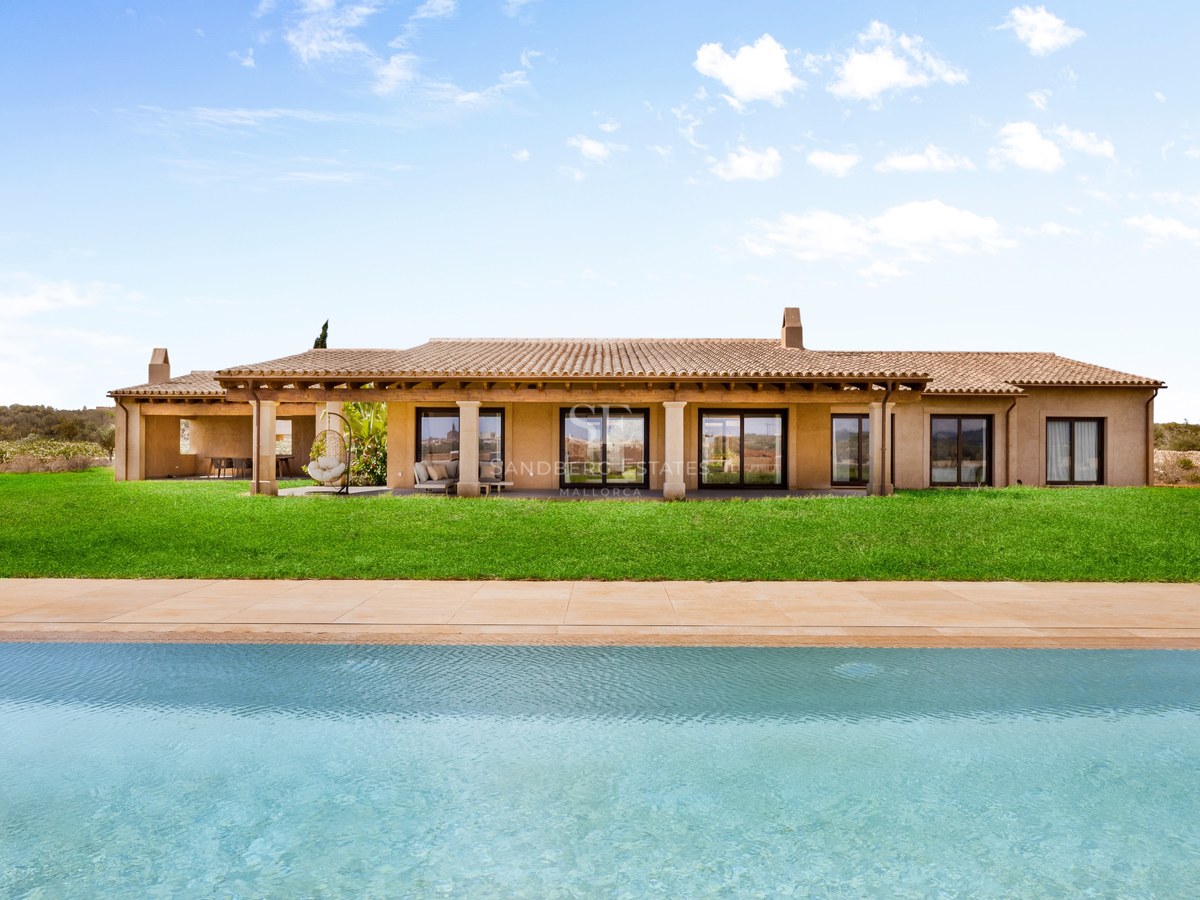 Large turquoise pool in front of a modern Mediterranean villa with a green lawn and terracotta roof under a blue sky.