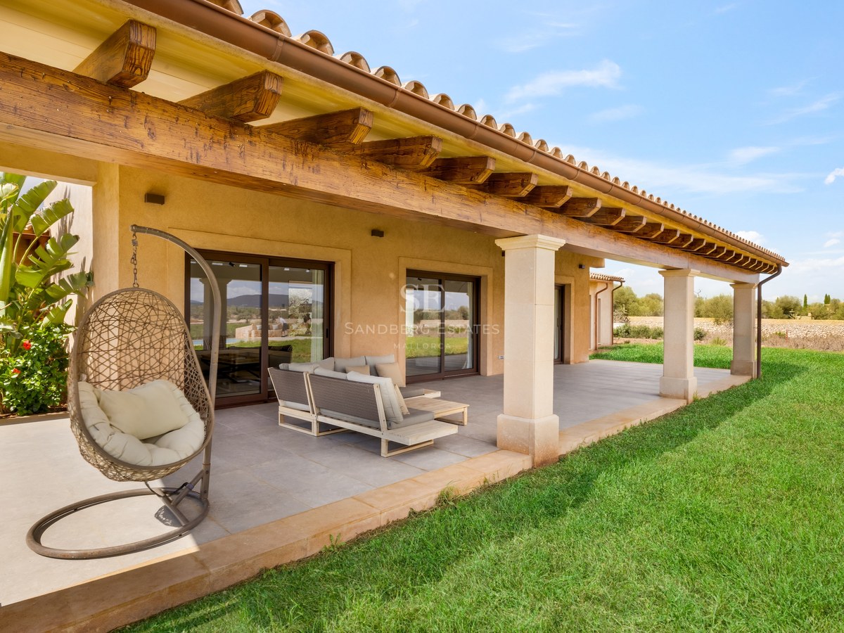 A covered terrace with rustic wooden beams, modern outdoor furniture, a hanging chair, and a green lawn under a blue sky.