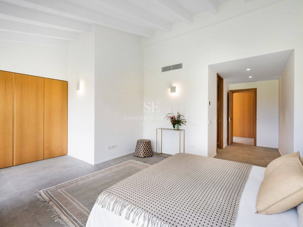 Bright bedroom with white beamed ceilings, wooden built-in wardrobes, large bed with beige pillows, and grey stone flooring.