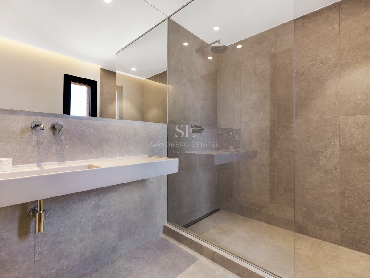 Minimalist bathroom with gray stone tiles, glass walk-in shower, wall-mounted sink, and large mirror.