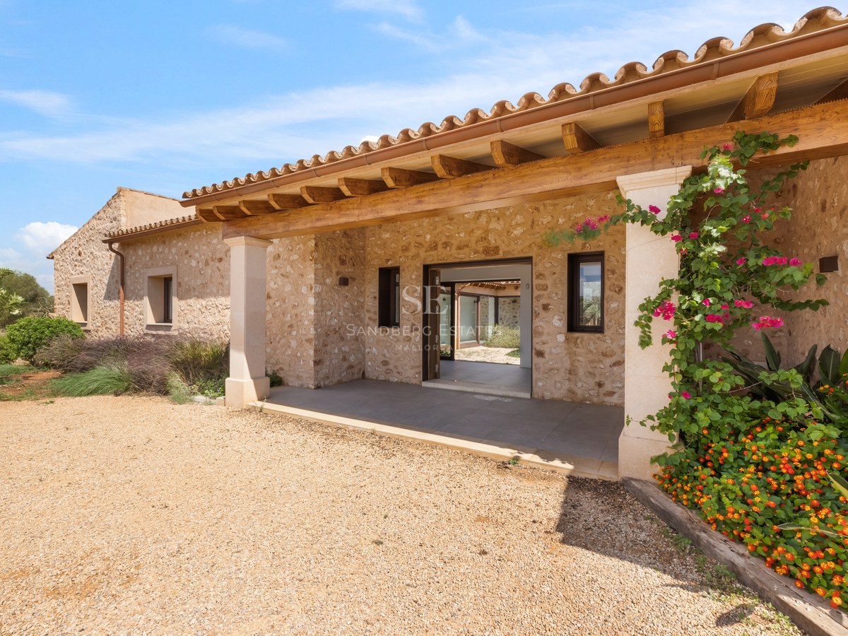 Entrance of a stone villa with a wooden porch, gravel path, and vibrant flowers under a blue sky.